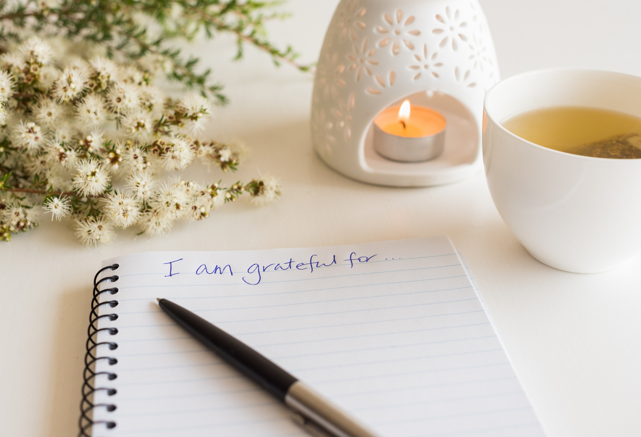 Close up of handwritten text "I am grateful for..." in foreground with notebook, pen, cup of tea, flowers and oil burner (Photo: iStock-natalie_board)