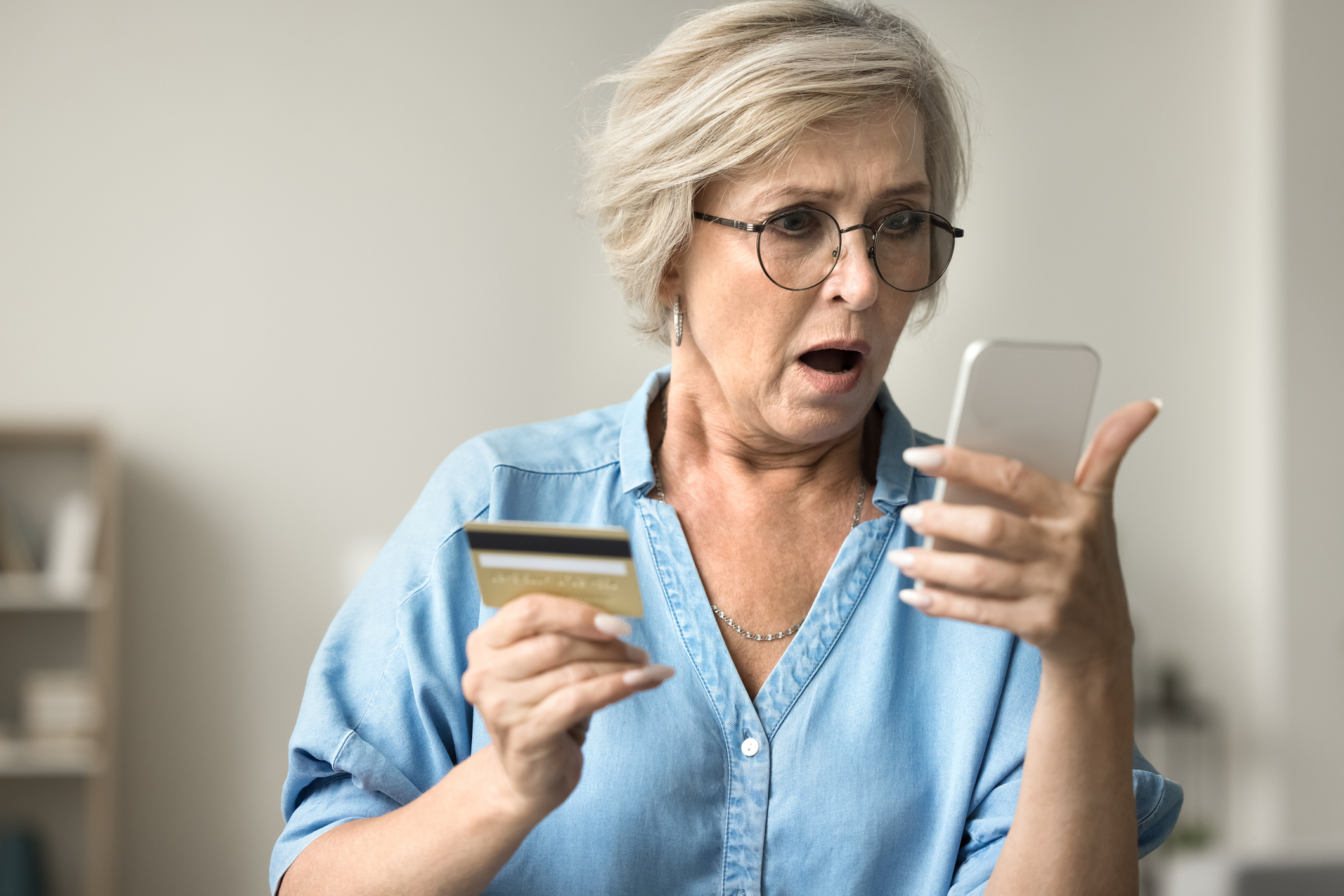Shocked concerned mature woman in glasses holding credit card, staring at smartphone in bad surprise. (Photo: iStock - fizkes)