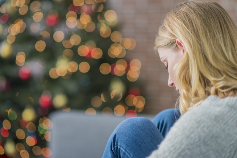 A woman is indoors in her living room. There is a Christmas tree in the background. The woman is wearing warm clothing. She is sitting on the couch and looking sad because she is alone on Christmas day. (Photo: iStock - FatCamera)