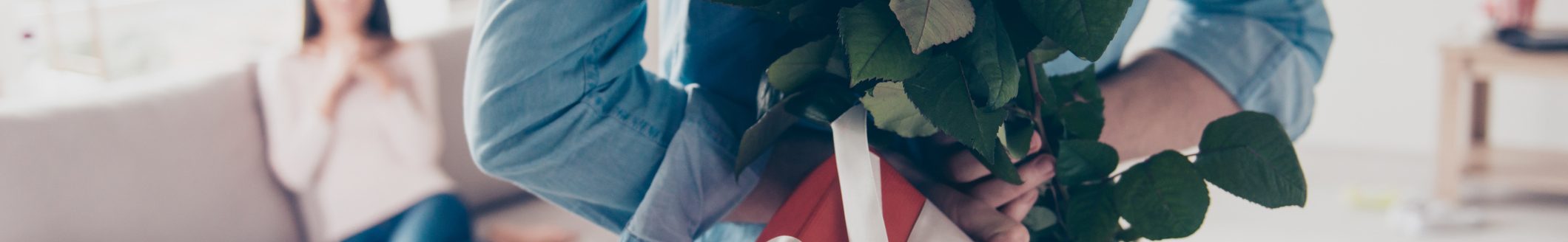 Man's hands hiding holding chic bouquet of red roses and gift with white ribbon behind back, happy woman is on blurred background (Photo: iStock - Deagreez)