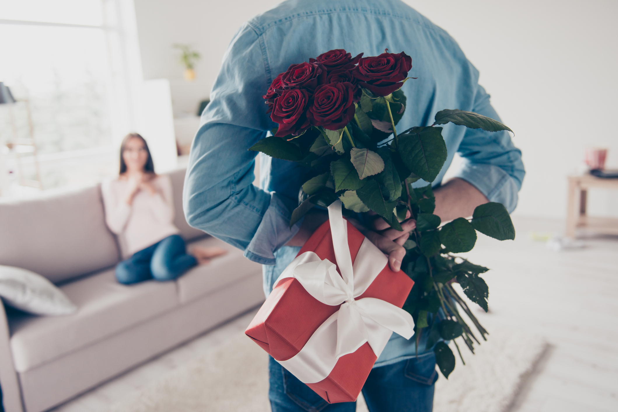 Man's hands hiding holding chic bouquet of red roses and gift with white ribbon behind back, happy woman is on blurred background (Photo: iStock - Deagreez)