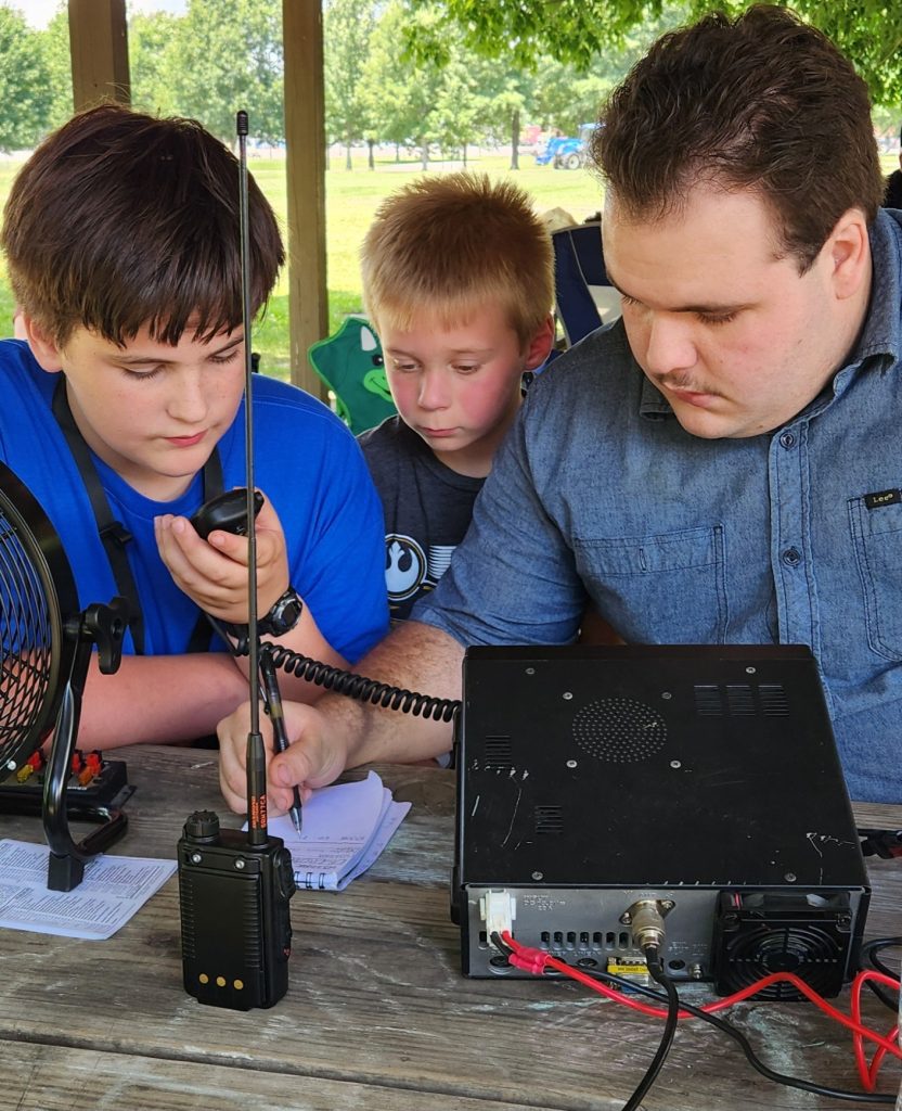 Oliver Baker, 6, of Metropolis, Illinois, takes a peek to see what Southernmost Illinois Emergency Radio Club members Drew Byrne (left), 14, of Kevil, Kentucky, and Aidan Carnes of Karnak, Illinois, are doing as they make a radio contact during ARRL Summer Field Day on Saturday. (Photo by Lloyd Baker)