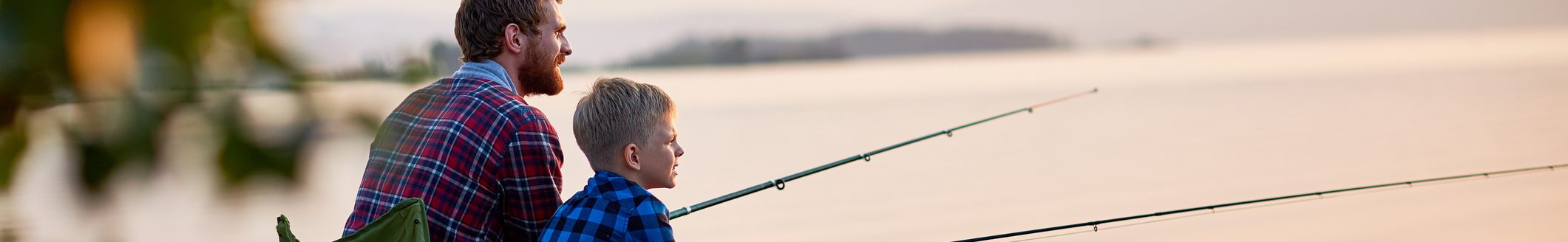 Father and son sitting together on rocks fishing with rods in calm lake waters with landscape of setting sun. (Photo: iStock │ #684090194 - shironosov)