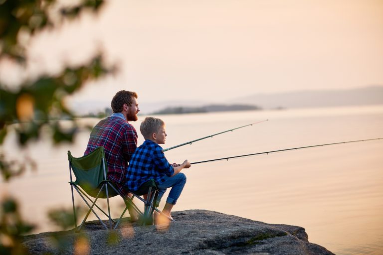 Father and son sitting together on rocks fishing with rods in calm lake waters with landscape of setting sun. (Photo: iStock │ #684090194 - shironosov)