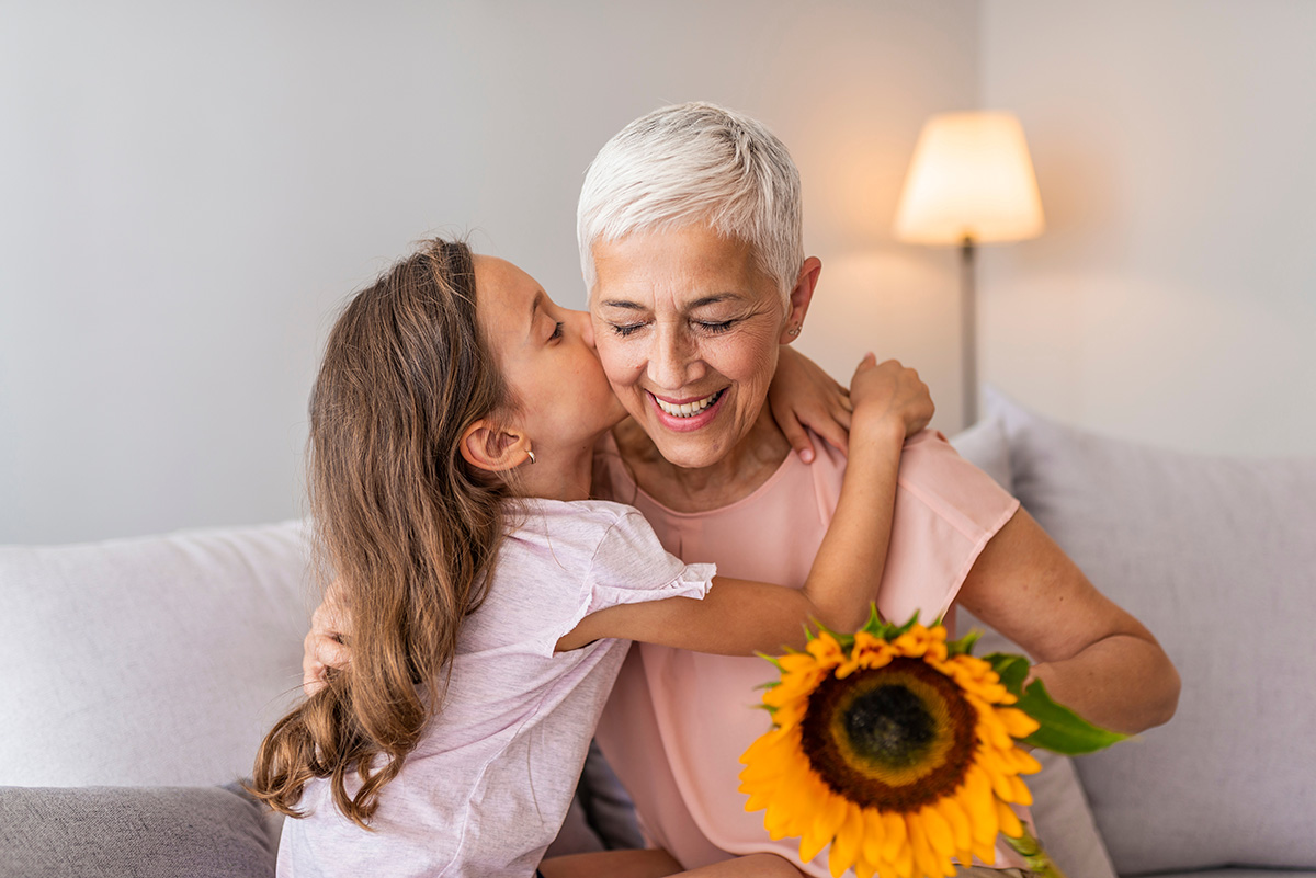 Little preschool granddaughter kissing happy older grandma on cheek, giving sunflower. (Photo: Adobe Stock │ #284777270 - Dragana Gordic)