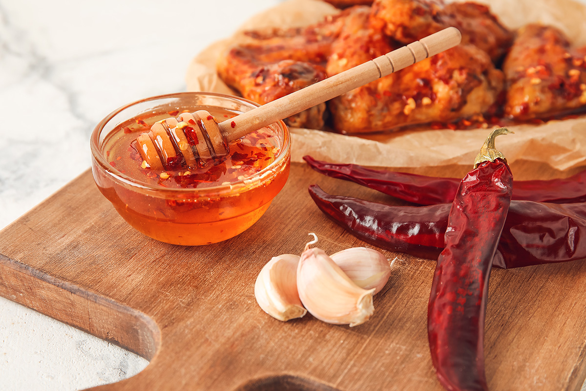 Bowl with hot honey, dry chili peppers, garlic and fried chicken wings on table (Photo: Adobe Stock │ #440769365 - Pixel-Shot)