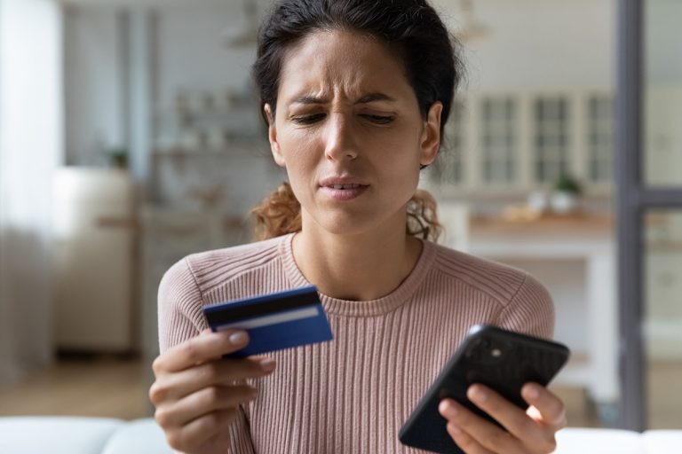 Latina woman holds credit card, smart phone. Victim of fraud. (Photo: Adobe Stock │ #482912516 - fizkes)