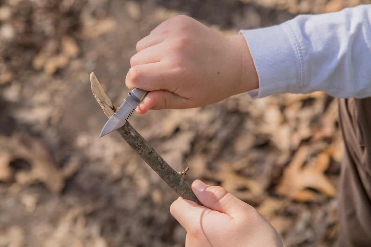 A young boy uses a knife to whittle a stick while out hiking in the wilderness. (Photo: iStock │ #469149404 - AmyKerk)