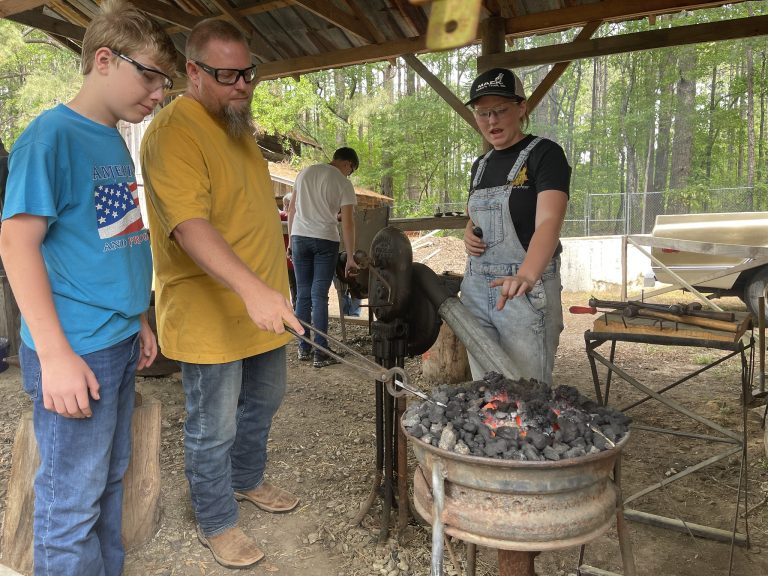 At a meeting of the Cleveland County 4-H Blacksmithing Club, held at the Pioneer Village in Rison, club member Millie Montgomery, 15, explains how club volunteer Jeff Frey and his son Brayden Frey, 13, are heating a piece of low-carbon steel to be hammered into a tent stake. (Division of Agriculture photo.)