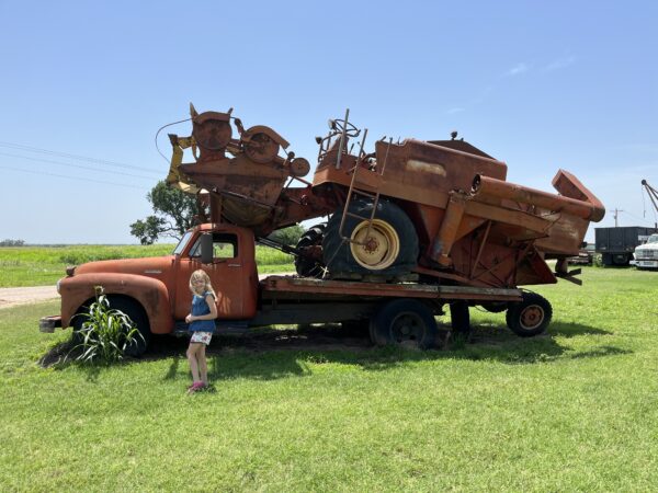 Zoey standing by an old Chevy with a rusty combine loaded on back. 