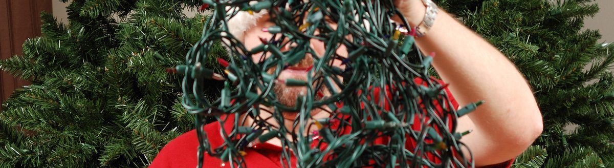 A man attempts to untangle a string of Christmas lights in front of an undecorated Christmas tree. (iStock │ #153787262 - MatthewSinger)