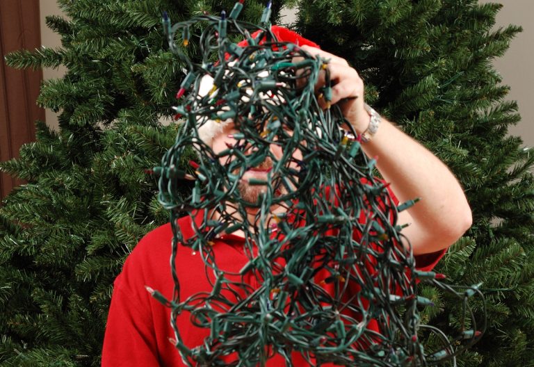 A man attempts to untangle a string of Christmas lights in front of an undecorated Christmas tree. (iStock │ #153787262 - MatthewSinger)