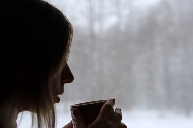 Long hair woman standing among the window and drinking hot tea from mug. Winter landscape. Lady drinks coffee looking at snowfall. Depression, seasonal affective disorder. (iStock │ #1940026348 - Marina Demidiuk)