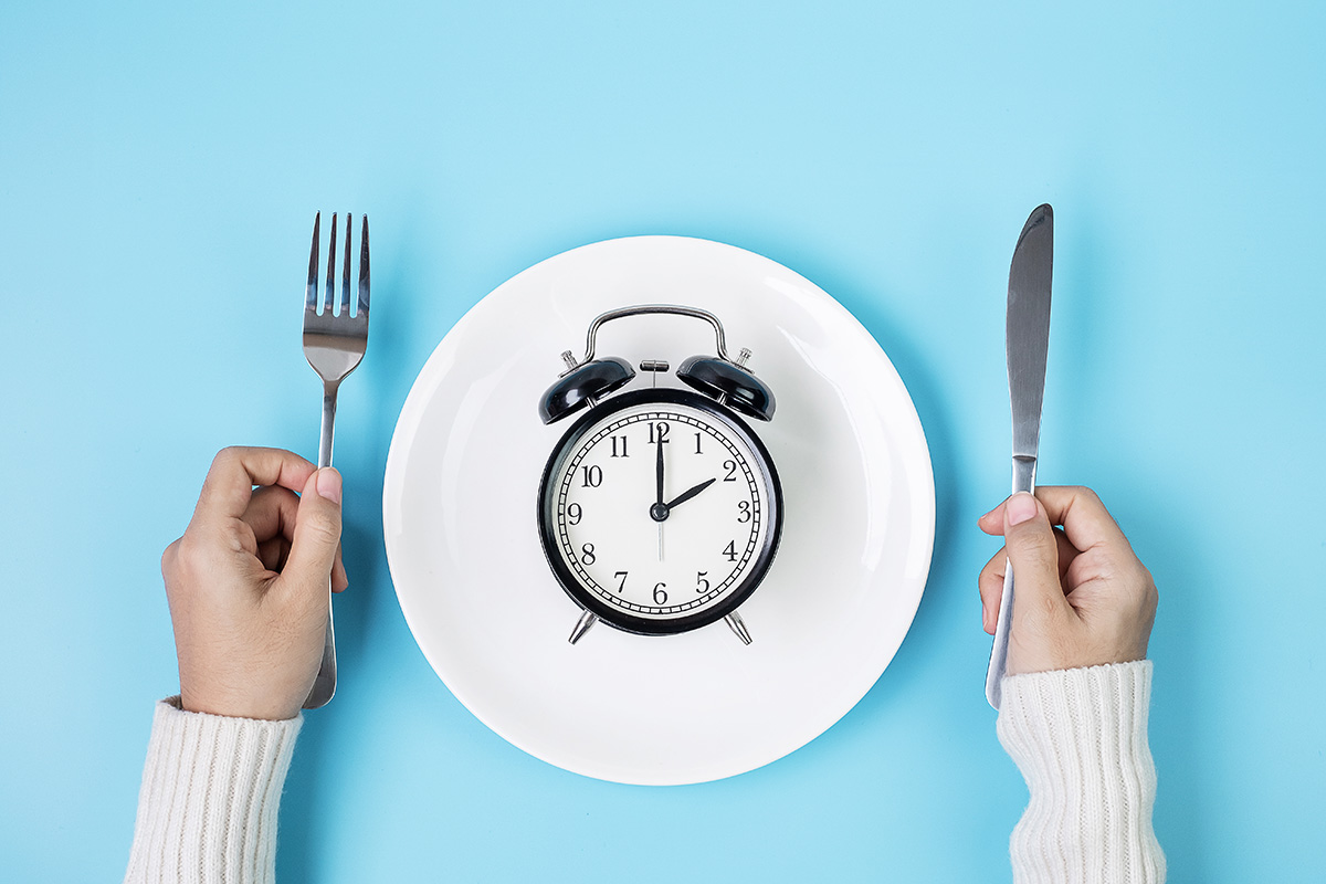 Hands holding knife and fork above alarm clock on white plate on blue background. Intermittent fasting. (Adobe Stock │ #389885849 - Jo Panuwat D)