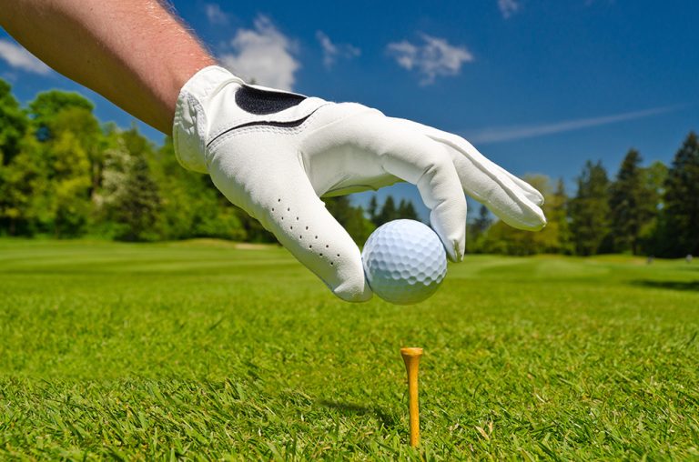 Hand placing golf ball on tee over beautiful golf course with blue sky. (Adobe Stock-#127222431 │ karamysh)