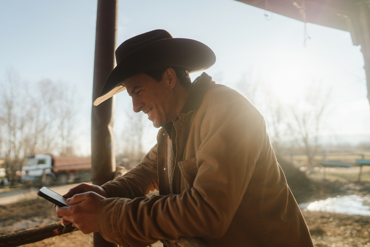 Adult man with a cowboy hat, using smart phone on his farm. (iStock-#2249520688 │ AleksandarNakic)