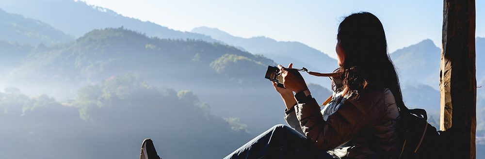 Woman traveler with camera enjoying a mountain view. (Adobe Stock-#1205421233 │ torjrtrx)