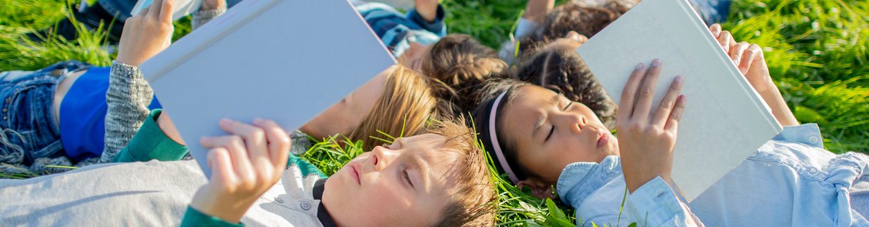 A small group of school-aged children lay in the grass on a sunny summers day as they each read a book. (iStock-#2164505455 │ FatCamera)