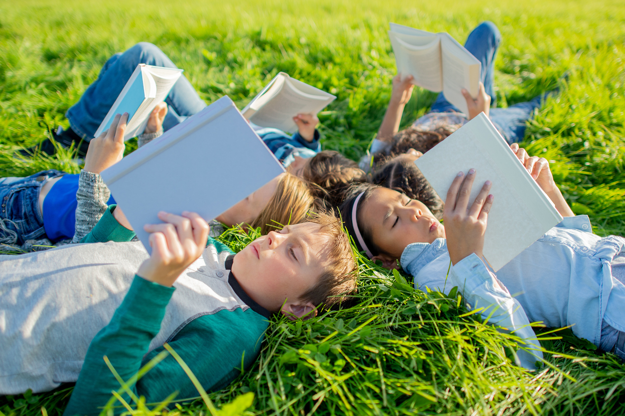A small group of school-aged children lay in the grass on a sunny summers day as they each read a book. (iStock-#2164505455 │ FatCamera)