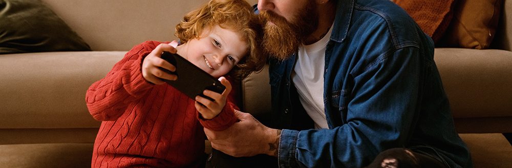 Father and son playing online game on mobile phone together while sitting on floor. (Adobe Stock-#553398947 │ Drobot Dean)