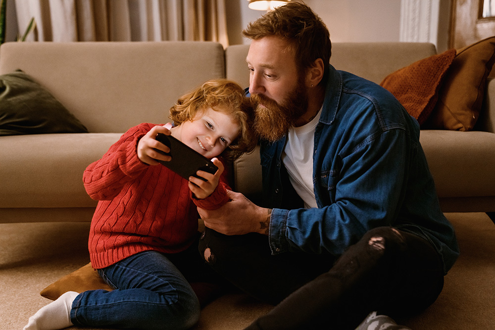 Father and son playing online game on mobile phone together while sitting on floor. (Adobe Stock-#553398947 │ Drobot Dean)