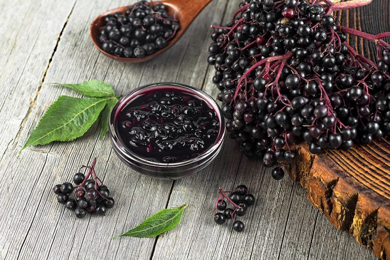 Homemade black elderberry syrup in glass jar and bunches of black elderberry with green leaves on wooden desk. (Adobe Stock-#625827026 │ domnitsky)