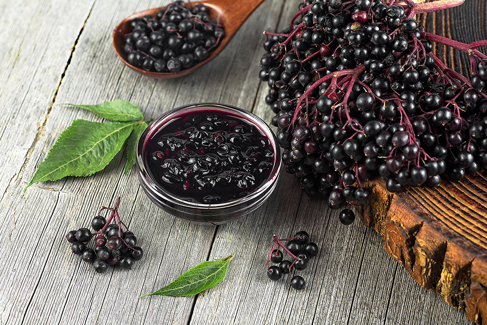 Homemade black elderberry syrup in glass jar and bunches of black elderberry with green leaves on wooden desk. (Adobe Stock-#625827026 │ domnitsky)